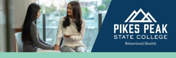 Two women are seated indoors, engaged in a supportive conversation. One woman is gently holding the other's hand while speaking. The Pikes Peak State College logo and the text 